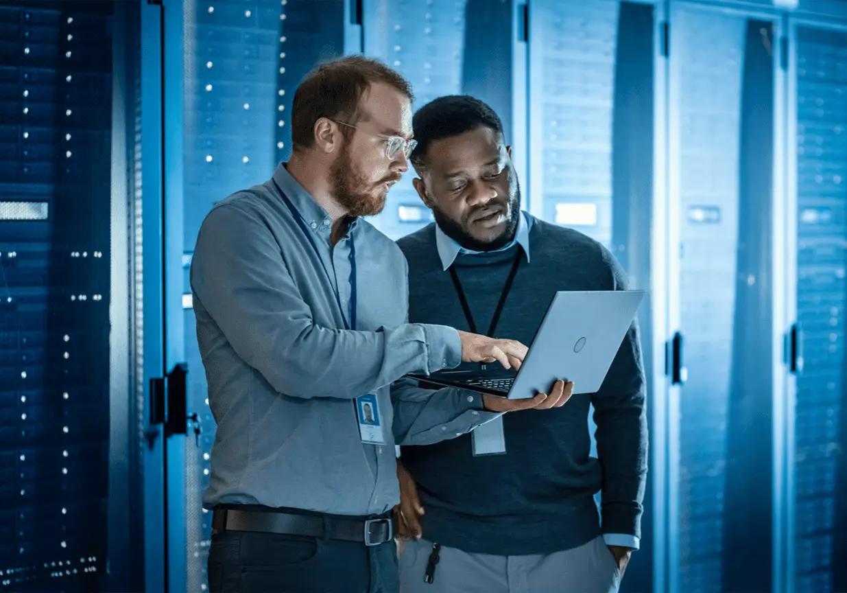 two men in server room one holding laptop and pointing into the screen